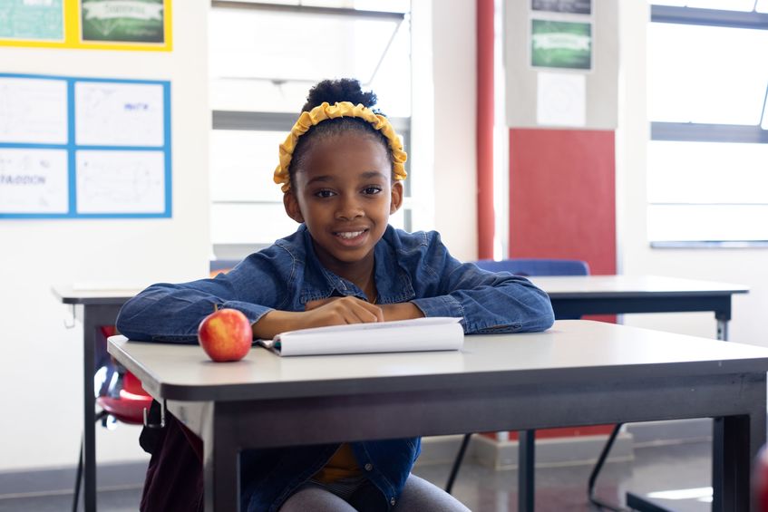 African American girl sitting at classroom desk with notebook, red apple, posters, window blinds. education, learning, student, study, academic, curriculum, school